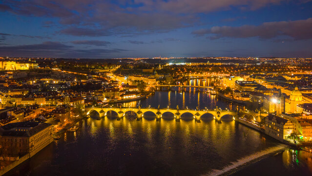 View of Charles Bridge in Prague at night. The bridge is illuminated and reflects in the river. Surrounding buildings are lit and the sky is darkening with clouds.