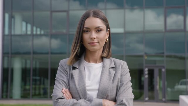 A confident and stylish young businesswoman stands with arms crossed in front of a modern glass corporate office building exterior smiling.