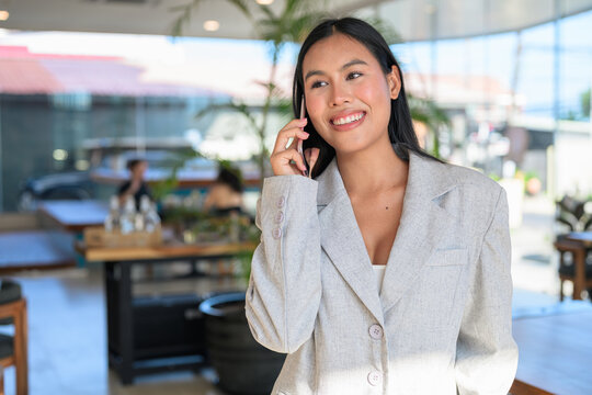 A smiling Asian woman from Thai Isan talking on a smartphone in a professional cafe.