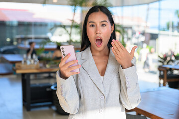 Fototapeta premium A shocked Asian woman from Thai Isan looking at a smartphone with an amazing expression in a professional cafe.
