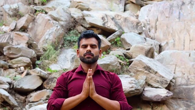 Young Indian man practicing meditation and prayer pose in nature, peaceful male meditating with closed eyes against rocky mountain background, spiritual wellness and mental health concept
