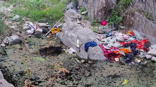 Environmental pollution with colorful old clothes and garbage dumped in a stagnant pond, water pollution and waste management issues in India
