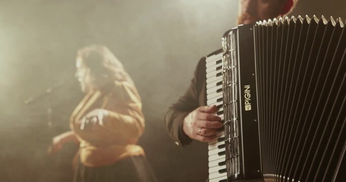 Bearded accordion player performing in warm smoky stage light with a blurred female singer in the background, creating a cinematic live music atmosphere