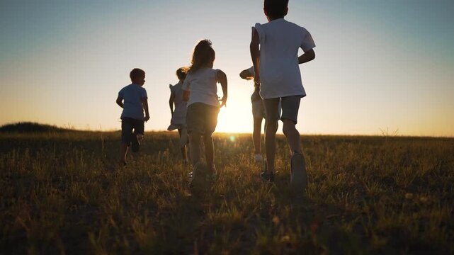 Group of children running across open field at sunset. Kids run together toward golden sun forming silhouette against sky. Child friendship and carefree freedom shine in warm summer sunlight.