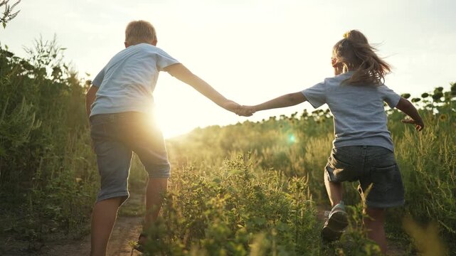 Boy and girl running together through beautiful nature. Happy child holding hand of sibling. Boy sprinting fast down long dirt path. Boy and girl running through summer field toward warm sunset.