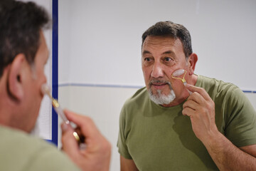 Mature man massaging face with jade roller in bathroom © Cristina Garcia