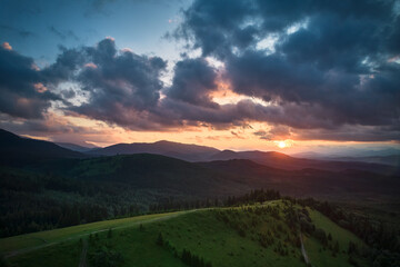 Obraz premium Aerial view breathtaking sunset over forest. Sun's rays break through dramatic clouds, casting golden glow across rolling hills and valleys. Distant mountains creating serene and picturesque scene.