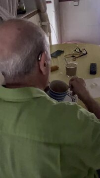 Desayuno de abuelo pensionista y jubilado con taza de caf&eacute;, sorbo y cuchara, sentado frente a televisi&oacute;n y aud&iacute;fonos en cocina de casa
