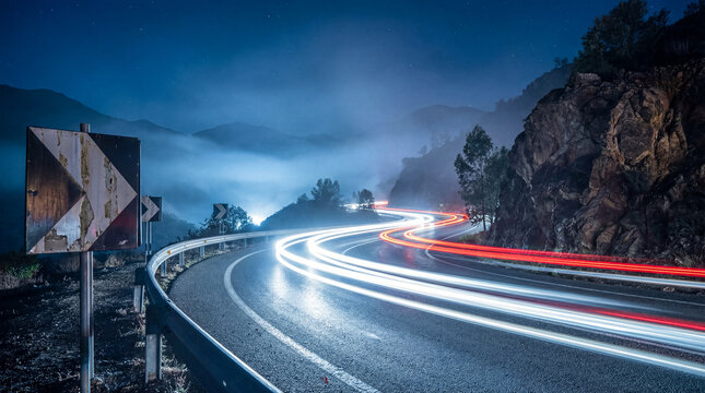 Long exposure photograph of car light trails on a winding mountain road at night, showcasing dynamic movement.