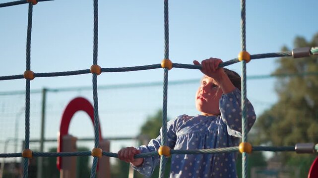 Little girl climbing rope net on playground. Child grips climbing rope and reaches up. Kid plays on outdoor playground equipment. Girl climbs net structure under clear blue sky on sunny day.