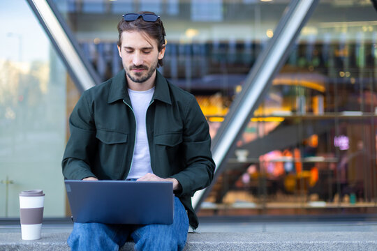 Freelancer working on laptop outdoors by modern glass office building