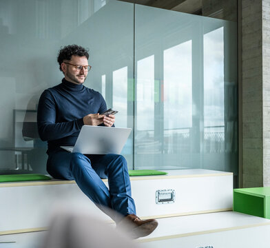 Businessman sitting in modern office smiling at mobile with notebook