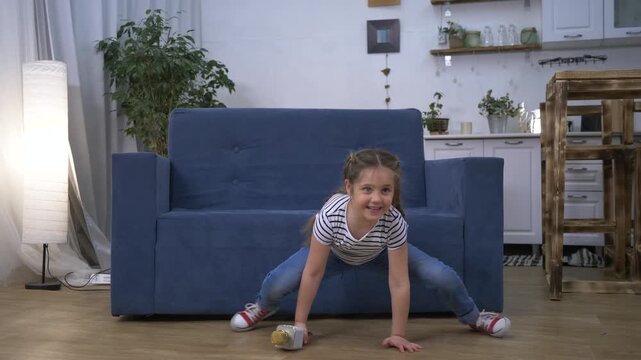 Happy young girl enjoys singing performance. Child holds toy microphone playing at home. Active girl does split on floor dancing. Happy singer holds microphone near blue sofa. Fun home play.