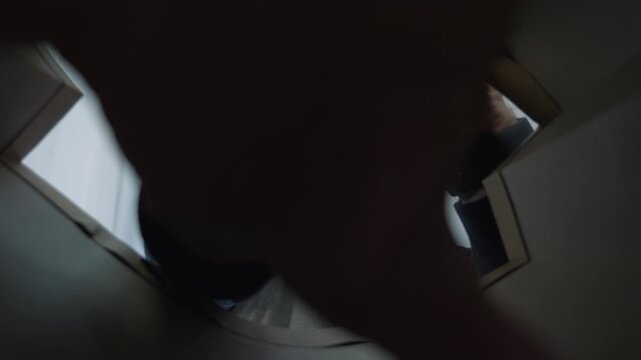 Man opening cardboard box viewed from inside. Person in face mask looks into parcel. Unboxing delivery package at home. Man peers into open box curiously. Package opening from interior perspective.