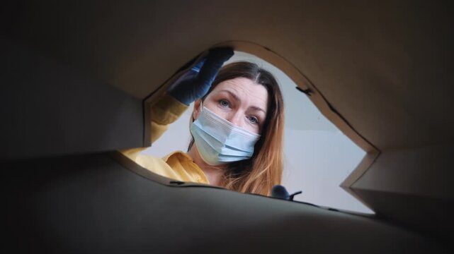 Woman opening cardboard box viewed from inside. She wears face mask and protective glove. Woman looks into package with curiosity. Delivery box unboxing from inside. Safe parcel delivery with mask on.
