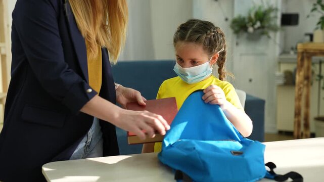 Mother helps child pack backpack for school. Girl in face mask holds blue bag open. Mom puts book into backpack at home. Child wearing mask prepares for school with parent helping at table.