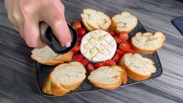 Woman in cozy kitchen attire prepares baked Camembert cheese, stuffing garlic slices into crisscross cuts on the rind, surrounding it with halved tomatoes and baguette chunks for oven roasting.