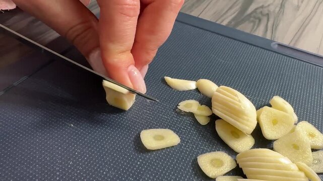 Close-up of woman chef slicing garlic into thin slices on wooden board.  Professional kitchen prep. Fresh clove, so very even thickness!