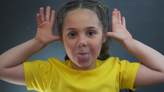 Funny child making silly face at camera. Little girl sticks tongue out and holds hands near ears. Playful kid makes funny grimace. Child teasing with funny expression. Girl enjoys being silly indoors.