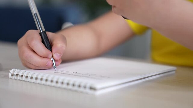 Child hand writing in notebook with pen. Kid writes notes on white paper page. Child doing homework at wooden desk. Hand holding pen and writing text. Student notebook with spiral binding on table.
