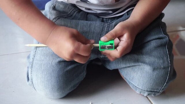 Close up of a little boy sitting on the floor, using a small green manual pencil sharpener to sharpen a wooden pencil.