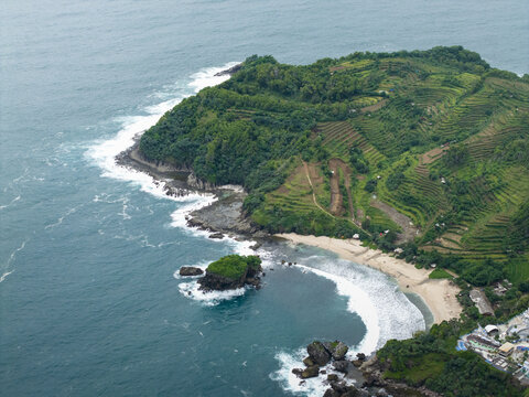 Aerial view of Jungwok Beach with lush green headlands, golden sands, deep blue ocean, and white waves, Gunungkidul Regency, Special Region of Yogyakarta, Indonesia.