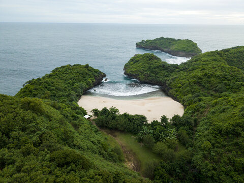 Aerial view of Sedahan Beach's secluded cove embraced by verdant cliffs meeting the turquoise sea under a soft sky, Gunungkidul Regency, Special Region of Yogyakarta, Indonesia.