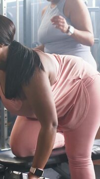 Vertical video: Kneeling on bench, pink woman rowing hex dumbbell to ribs, gray trainer cueing form