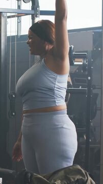 Vertical video: Starting warmup, woman in blue set raising and rotating arms by cable, duffel bag