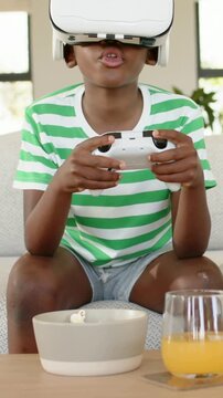 Vertical video: Playing boy in shirt pressing controller on sofa as headset showing, popcorn, juice