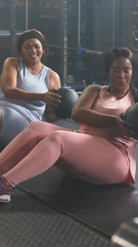 Vertical video: Cueing two women in blue and pink twisting passing medicine ball in gym for core