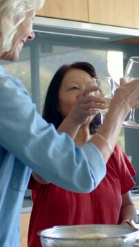 Vertical video: Reaching blonde in denim shirt and Asian in red top toasting with wine in kitchen