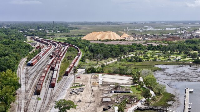 Aerial view of a railway yard with freight cars, green forests, a golden industrial pile, and a calm estuary, Fernandina Beach, Florida, United States.