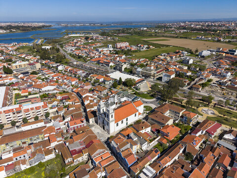 Aerial view of the striking white church contrasting against the red-tiled roofs of the buildings, framed by the blue river and distant horizon, Vila Franca de Xira, Portugal.