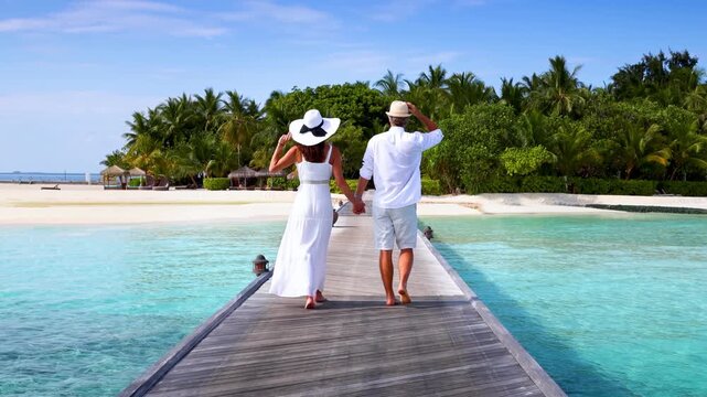 A elegant couple walks down a wooden pier over turquoise sea in the Maldives islands during their romantic holiday time