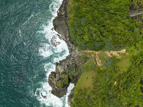 Aerial view of the rugged coastline where turquoise waters crash against dark rocks, contrasting with the vibrant green vegetation, Gunungkidul Regency, Daerah Istimewa Yogyakarta, Indonesia.