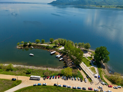 Aerial view of Lake Kerkini with boats docked at a small pier, lush green trees along the shoreline, and a parking area with cars under a clear blue sky in Serres, Greece.
