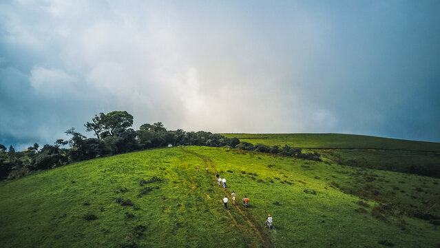 Aerial view of vibrant emerald slopes ascending towards a brooding, stormy sky, where tiny figures brave the winding path, Obudu, Cross River, Nigeria.