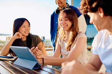 Fototapeta premium Group of smiling university students collaborating on a digital tablet at outdoor campus, fostering teamwork and learning