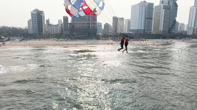 Two people are parasailing high above the sparkling ocean waves, enjoying exciting water sports and a panoramic view of the bustling city skyline and sandy beach filled with sunbathers below