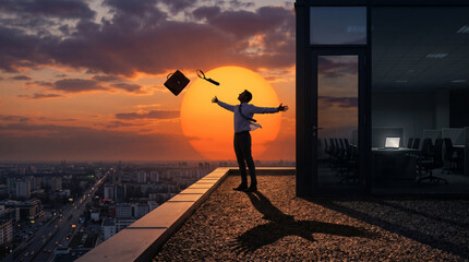 Plakat Businessman celebrating on rooftop overlooking city at sunset with briefcase in air.