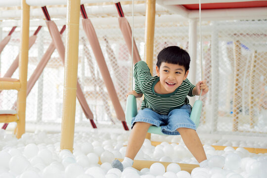 Active Asian kid on a pastel green swing, surrounded by white ball pits.