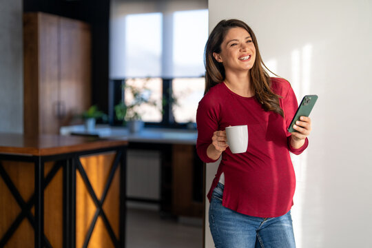 Beautiful happy fit pregnant woman talking on her phone while having a cup of coffee in the morning