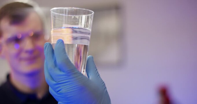A young man in safety glasses and blue gloves holding a glass of water while looking at it in a laboratory