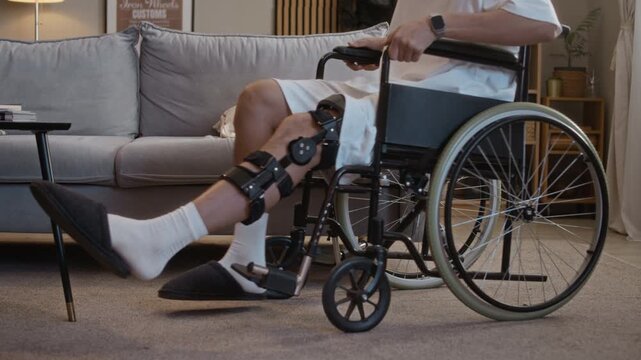 Medium shot of anonymous young man with orthopedic brace on leg standing up and getting into wheelchair at home