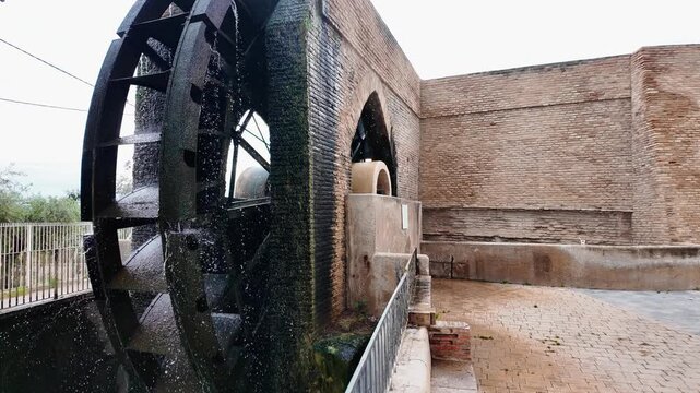 Historical water wheel standing against a weathered stone wall structure of an ancient hydraulic mill with metal railings under soft daylight in a rural outdoor setting showcasing traditional engineer