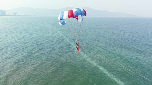 Person parasailing over ocean waters, enjoying an adventurous water sport activity with a city skyline and beach in the background on a sunny day, experiencing freedom and excitement