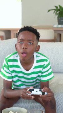Vertical video: Gaming boy in striped tee reacting to game on sofa holding gamepad eating popcorn