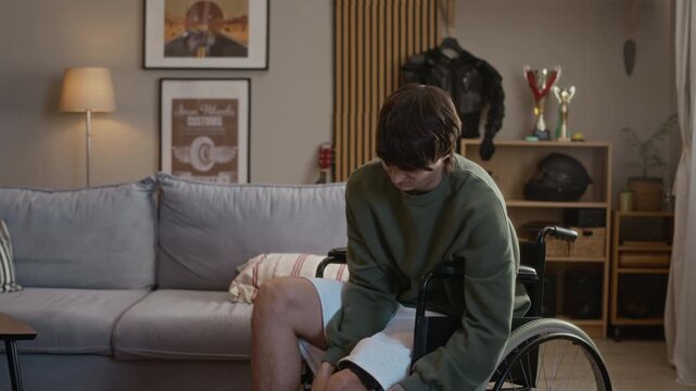 Tilt up shot of young Caucasian man sitting in wheelchair and adjusting orthopedic brace, wincing in pain and suffering