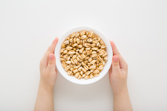 Little child boy hands holding and showing full white bowl of yellow dry puffed wheat flakes on light gray table background. Flat lay. Closeup. Point of view shot. Top down view.